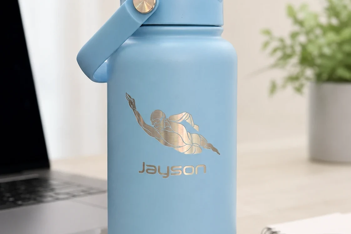 Sky Blue BE FIT SWIGGER motivatie waterfles met Jayson naamgravering en zwemmer illustratie op een lichte werkplek.